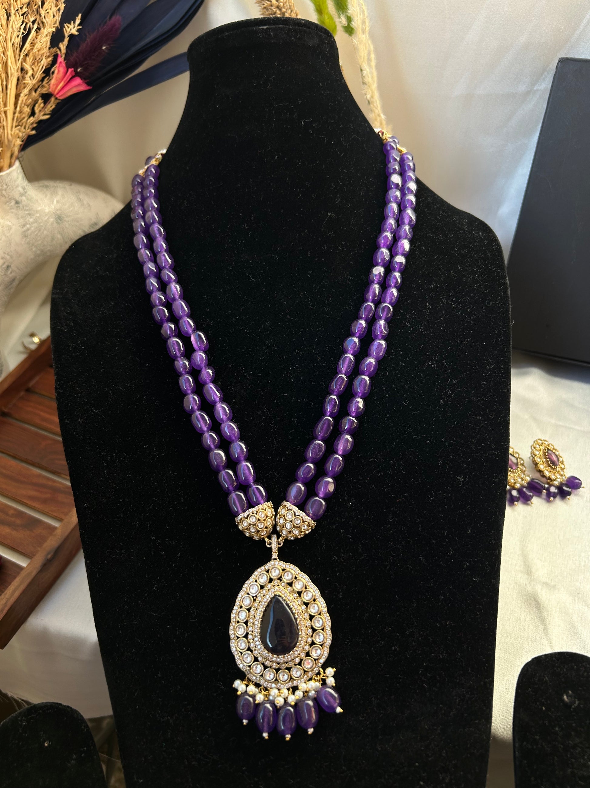 Parineeti purple Kundan Rani Haar – A gold-plated Rani Haar necklace with purple gemstones and matching earrings, set on a rustic wooden backdrop."
