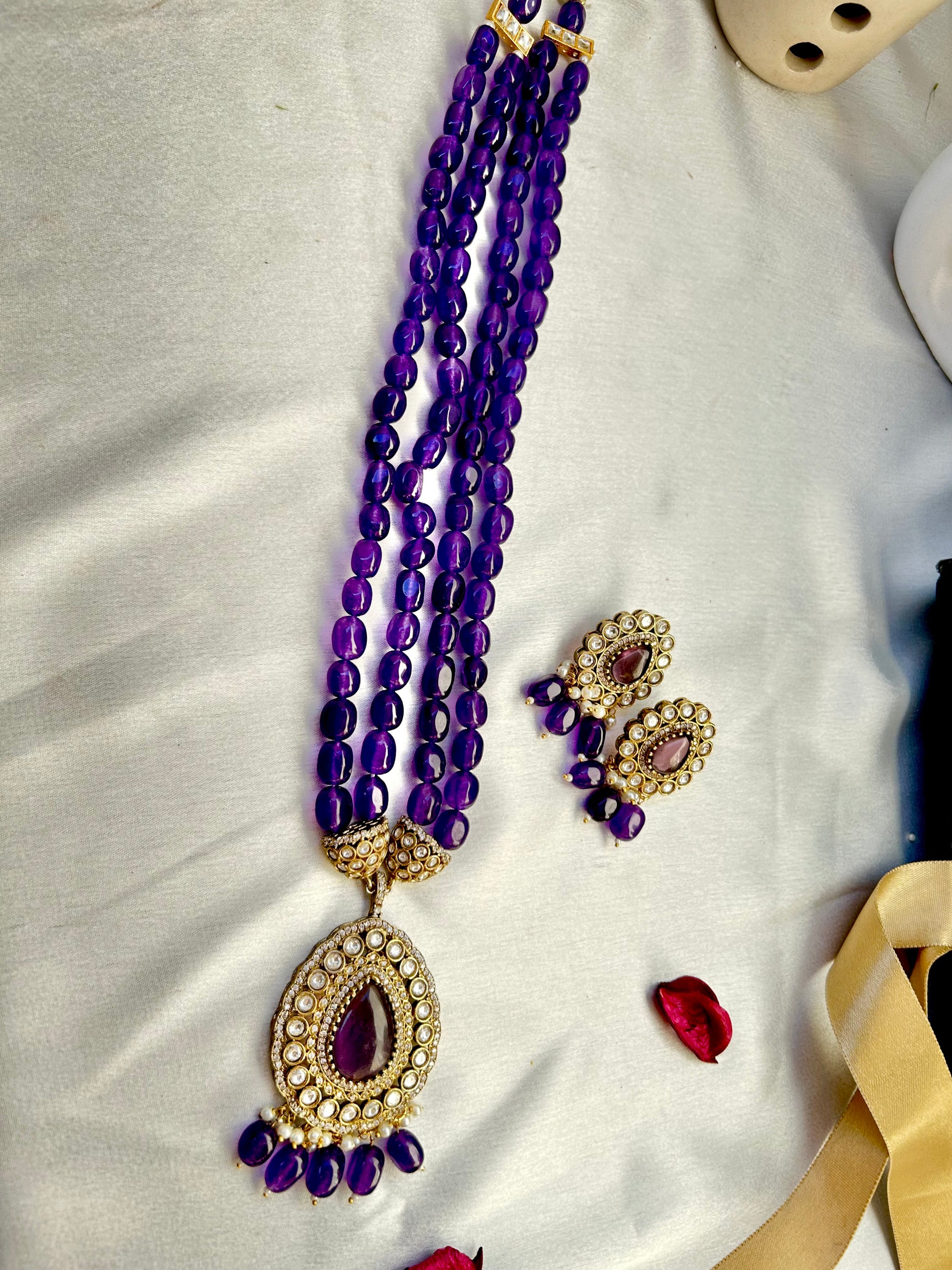 Parineeti purple Kundan Rani Haar – A gold-plated Rani Haar necklace with purple gemstones and matching earrings, set on a rustic wooden backdrop."